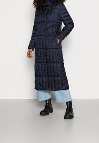 Abrigo largo de pluma de color azul marino con patrón de acolchado vertical, cierre completo, bolsillos y capucha ajustable. Lleva puesto un pantalón de mezclilla ancha de color azul claro y botas negras.