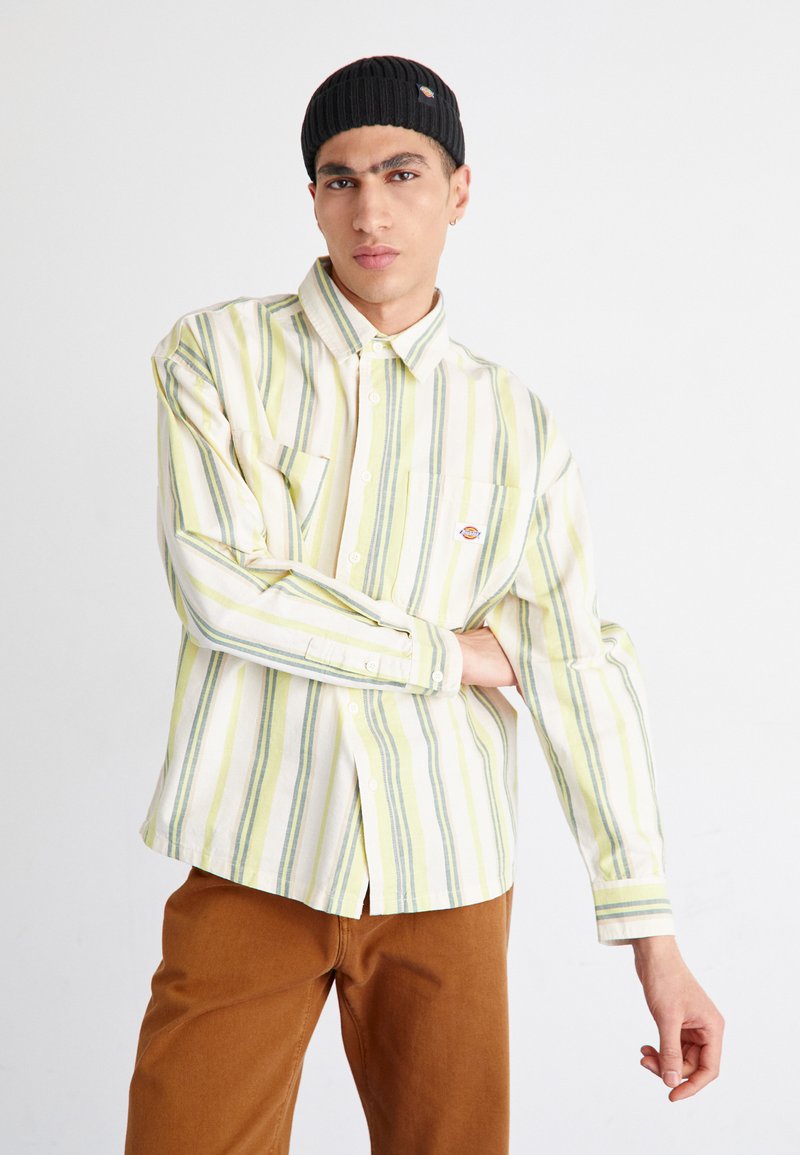 Camisa de botones a rayas en colores amarillo, verde y blanco, con un bolsillo en el pecho y un logo redondo. Combinada con pantalones marrones.