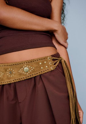 Persona che indossa una canottiera marrone e pantaloni con una larga cintura in camoscio beige con borchie metalliche e lunghe frange legate in vita.