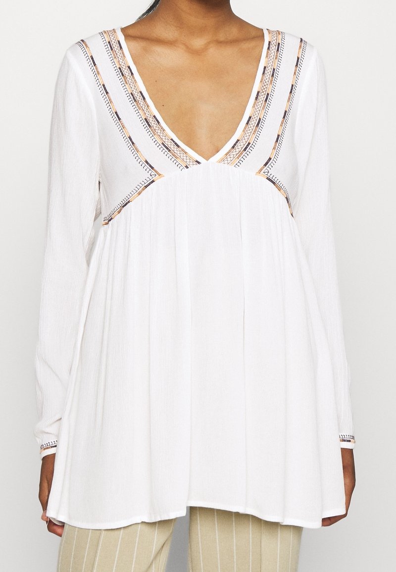 Blouse blanche à manches longues avec un col en V profond et un galon brodé multicolore portée avec un pantalon clair à rayures.
