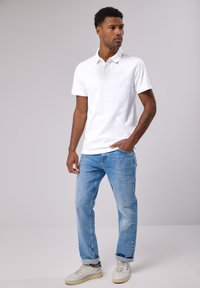 Bílá bavlněná polo košile s límečkem, doplněná světle modrými džínami. Oblečení obsahuje bílé tenisky s zelenými akcenty.