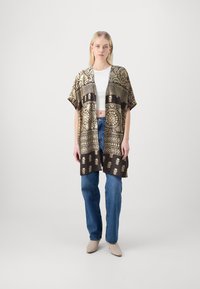 Kimono-stil yttertøy i svart og gullmønstret stoff, med løse ermer og avslappet passform, brukt over en hvit topp og jeans.