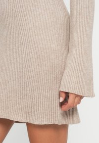 Vestido de punto acanalado beige con mangas largas y acampanadas, mostrado desde la mitad del muslo hasta la mano con un tono de piel natural y esmalte de uñas rosa claro.