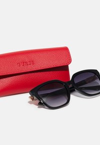 Black oversized sunglasses with gradient lenses next to a red textured leather case embossed with "GUESS."