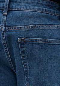 Κοντινή λήψη στην πίσω τσέπη και τις λεπτομέρειες της ραφής σε σκούρο μπλε denim τζιν με εμφανείς πορτοκαλί ραφές.