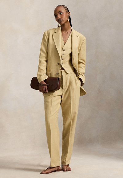 Ανοιχτό κίτρινο κοστούμι με σακάκι ραμμένο στα μέτρα, εφαρμοστό γιλέκο και παντελόνι με ίσια γραμμή. Κρατάει καφέ φάκελο και φοράει ταμπά σανδάλια.