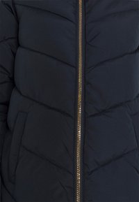 Meripuku takki, jossa on chevron-kviltattu muotoilu, kultaisella vetoketjulla ja sivutaskuilla. Pehmeä, sileä rakenne.