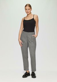Graue, maßgeschneiderte Hose mit Falten und Seitentaschen, kombiniert mit einem schwarzen Top mit Spaghettiträgern. Das Model steht mit den Händen in den Taschen. Schwarze Schuhe.
