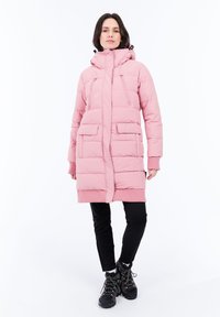 Una mujer se encuentra de pie vistiendo un largo abrigo acolchado rosa con capucha, pantalones negros y botas de senderismo negras, todo ello contra un fondo blanco.