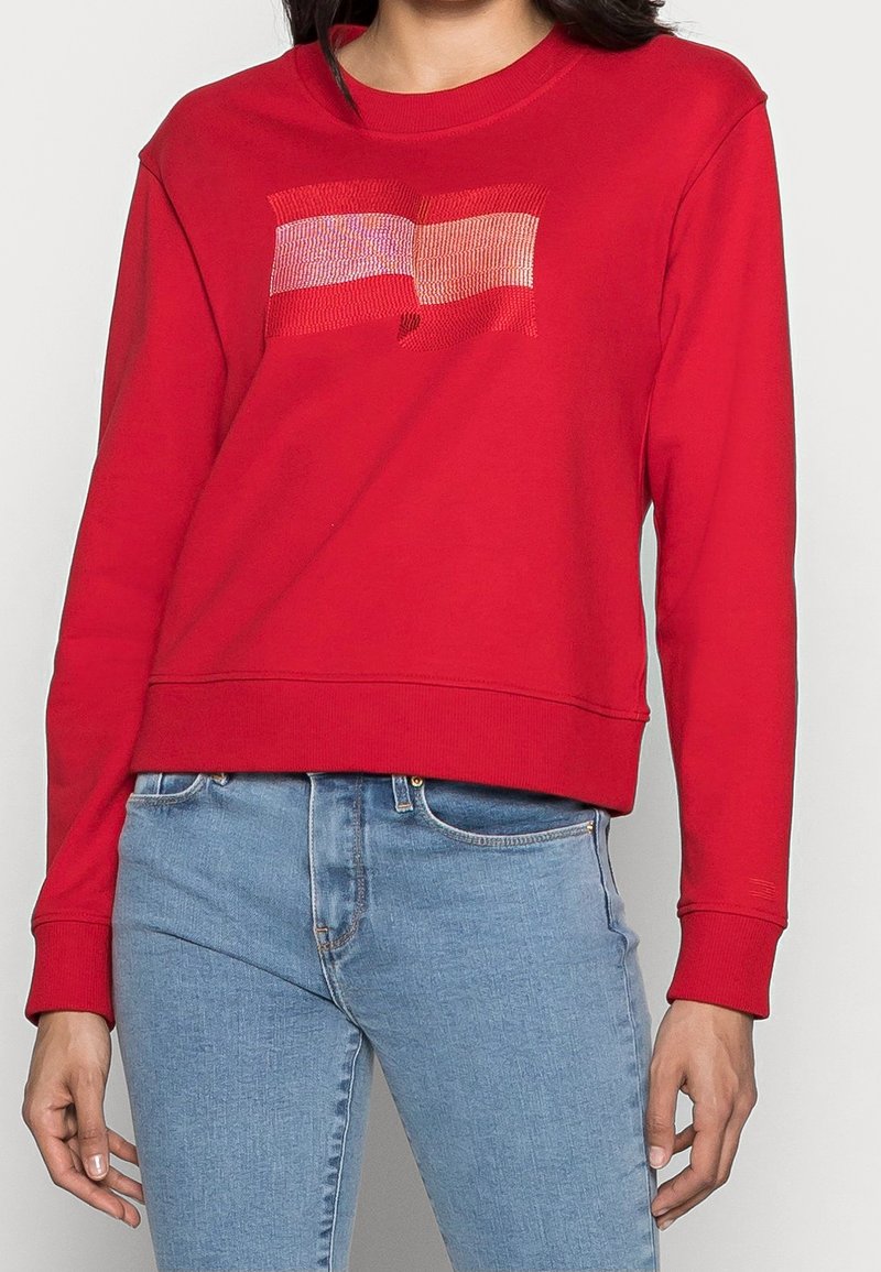 Personne portant un sweat-shirt rouge à manches longues avec deux drapeaux rectangulaires brodés sur la poitrine et un jean bleu clair, debout sur fond blanc.