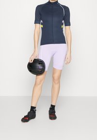 Maillot de cyclisme bleu marine à manches courtes avec des accents réfléchissants, assorti à un short violet clair. Porte un casque noir et des chaussures de cyclisme.