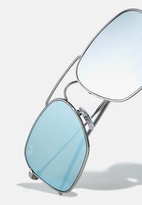 Ochelari de soare supradimensionați din metal cu lentile gradient albastre, având un cadru argintiu și tampoane de nas transparente, evidențiind o formă geometrică distinctivă.