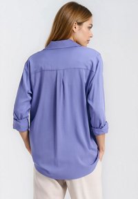 Jasnofioletowa koszula na guziki z podwiniętymi rękawami, miękka w dotyku, z tylnym zakładką i zaokrąglonym dołem, pokazana od tyłu, w połączeniu z jasnymi spodniami.