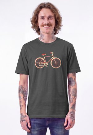 Junger Mann mit Tattoos und Nasenring lächelt, trägt dunkles T-Shirt mit Fahrradgrafik und blaue Jeans vor weißem Hintergrund.