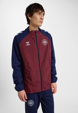 Junger Mann, der eine bordeauxrote und marineblaue Sportjacke und Hose mit Logos des Dänischen Fußballverbands trägt, steht da und schaut zur Seite.