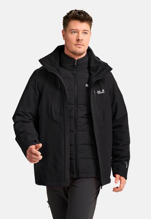 Man wearing layered black Jack Wolfskin outdoor jackets, standing and looking slightly to the side against a plain background.
