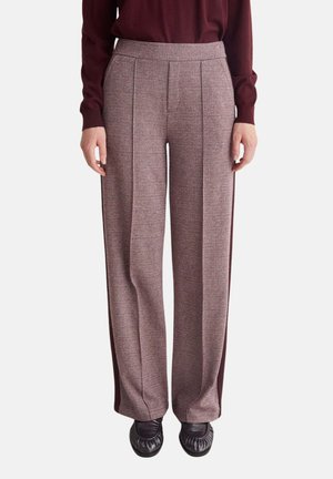 Persona que lleva pantalones de tiro alto y perneras anchas con franjas laterales burdeos, combinados con zapatos marrón oscuro y una camiseta de manga larga burdeos.