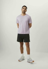 Man staand met handen achter zijn rug, gekleed in een lavendelkleurig t-shirt, zwarte shorts, witte sokken en zilveren hardloopschoenen, op een effen achtergrond.
