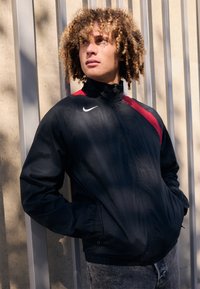 Veste coupe-vent noire avec des accents rouges, col haut et fermeture éclair. Portée par-dessus un jean gris, avec un logo Nike sur la poitrine.