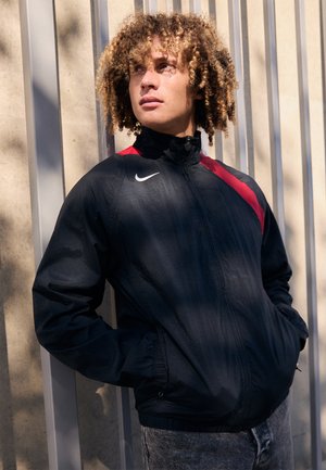 Veste coupe-vent noire avec des accents rouges, col haut et fermeture éclair. Portée par-dessus un jean gris, avec un logo Nike sur la poitrine.
