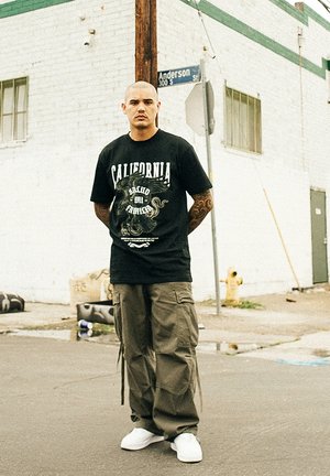 Sort grafisk t-shirt med "California" tekst og blæksprutte-design, parret med olivenfarvede cargo bukser og hvide sneakers, sat på en gadebaggrund.