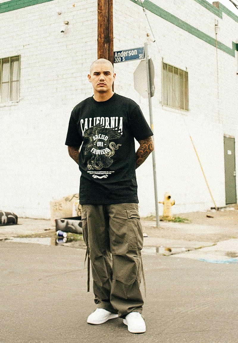 Sort grafisk t-shirt med "California" tekst og blæksprutte-design, parret med olivenfarvede cargo bukser og hvide sneakers, sat på en gadebaggrund.