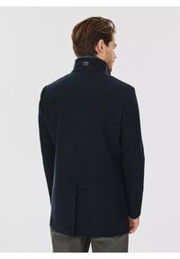 Granatowy płaszcz z wełny z domieszką, z wysokim kołnierzem, fakturowaną powierzchnią i pojedynczym rozcięciem z tyłu. Posiada małe logo w pobliżu kołnierza.