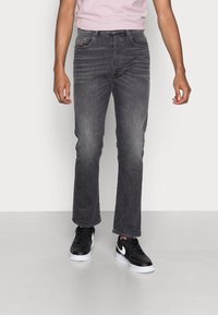 Jeans de mezclilla gris con corte recto, sutil desgastado, diseño de cinco bolsillos y un logotipo bordado en el bolsillo frontal. Llevados con zapatillas negras.