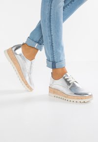 Chaussures métalliques argentées avec un design épuré, bout rond et semelle en liège texturée. Lacets blancs et semelle extérieure striée contrastante.