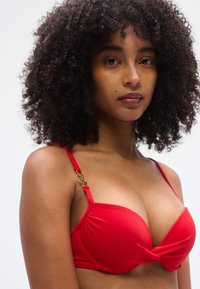 Κόκκινο μαλακό σουτιέν με λεπτομέρεια κόμπου στο κέντρο, ρυθμιζόμενους ιμάντες και διακοσμητικά στοιχεία σε χρυσή απόχρωση. Ομαλή, ελαστική υφή υφάσματος.