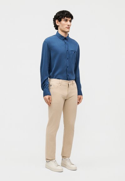 Άνδρας στέκεται φορώντας μπλε πουκάμισο με μακριά μανίκια και κουμπιά, μπεζ παντελόνια και άσπρα αθλητικά παπούτσια, μπροστά σε απλό ανοιχτό φόντο.