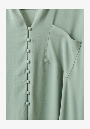 Chemise en tissu vert clair avec des boutons recouverts de tissu sur le devant et une seule poche poitrine du côté droit.