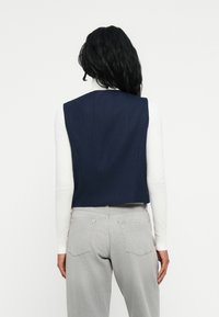 Kobieta z długimi czarnymi włosami, ubrana w granatową kamizelkę bez rękawów na biały golf i jasnoszare dżinsy, zwrócona plecami na jednolitym tle.