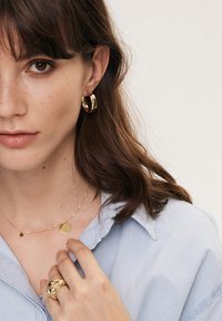 Złote kolczyki w kształcie kół, delikatny złoty naszyjnik z okrągłym wisiorkiem oraz teksturowany złoty pierścionek. Widoczna jasnoniebieska koszula z kołnierzykiem.