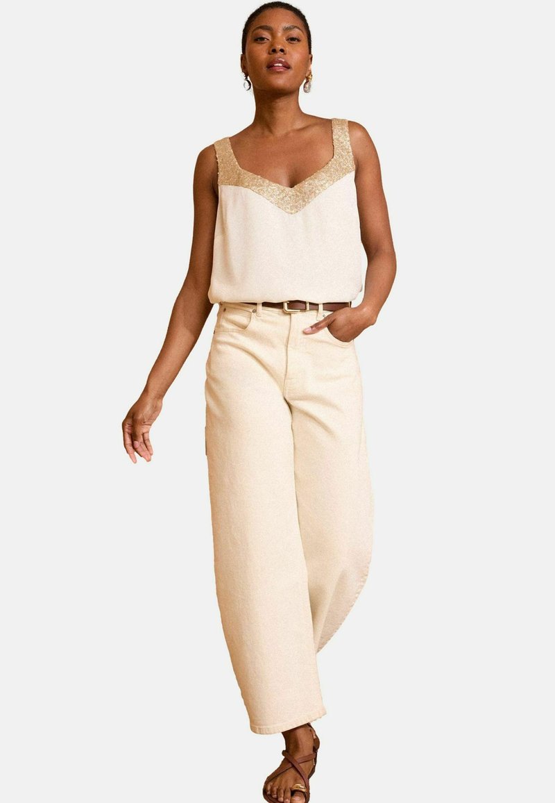 Beige sleeveless top with decorative gold accents, paired with high-waisted, wide-leg beige trousers. Notable belt detail and strappy sandals.