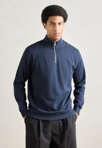 Námornícky modrý pulóver na zips z mäkkej tkaniny, ktorý má vysoký golier, dlhé rukávy a rebrované manžety a lem. Nosený s čiernymi nohavicami.