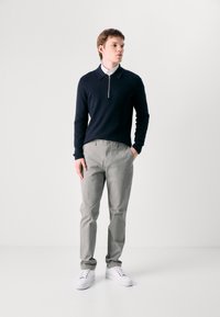 Marineblå genser med glidelås i kragen, grå bukser og hvite joggesko. Genseren er ribbete; buksene har en glatt tekstur. Minimalistisk design.