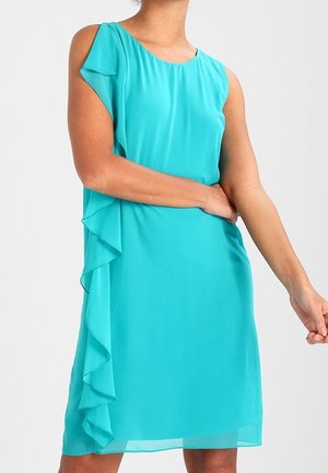 Mujer con vestido turquesa sin mangas con detalle de volantes a lo largo del lado izquierdo sobre un fondo claro y liso.
