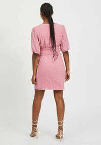 Rosa, texturerad klänning med puffärmar och elastisk midja. Matchas med beige remsandaler. Bakgrunden är neutral och enkel.