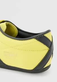 Sapatos Adidas Tokyo com um cabedal em tecido amarelo brilhante, detalhes em preto, costuras e um design elegante com perfil baixo.