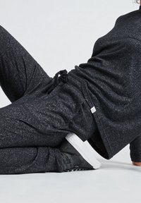 Persoon in donkergrijze joggingsbroek en sneakers, leunend op één arm tegen een effen lichte achtergrond.