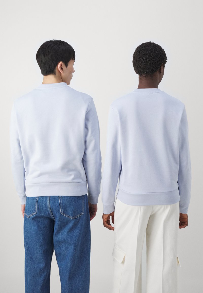 Lichtblauwe sweatshirts met lange mouwen, geribbelde manchetten en zoom, gedragen door twee personen in denim- en beige broeken, met hun rug naar de camera gekeerd.