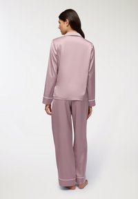 Set di pigiama rosa satinato con maniche lunghe e pantaloni a gamba larga. Presenta piping bianco a contrasto lungo i polsini e il colletto. Texture liscia.