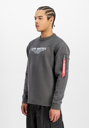 Young man with braided hair wearing a gray Alpha Industries sweater with text and a red "Remove Before Flight" tag on the sleeve.