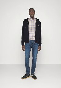 Camisola com fecho de correr azul-marinho sobre uma camisa de padrão às riscas, jeans de ganga azul, sapatos pretos com detalhes brancos e acabamento texturado. Roupa simples e casual.