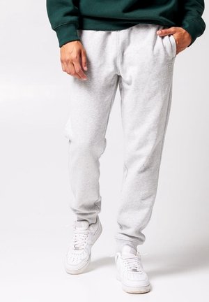 Mand iført lysegrå joggingbukser og hvide sneakers, med den ene hånd i lommen og den anden afslappet ned langs siden.