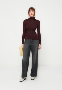 Burgunderfarbener, gerippter Rollkragenpullover, kombiniert mit weit geschnittenen, dunkelgrauen Jeans. Hält eine gelbe Clutch. Cremfarbene Stiefeletten mit einer kontrastierenden schwarzen Sohle.