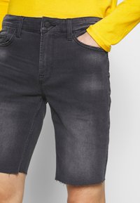 Tummanharmaat denim-shortsit, joissa on revitty helma, etutaskut ja napitus. Yhdistetty keltaiseen pitkähihaiseen paitaan.