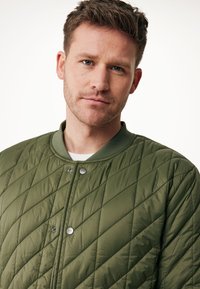 Prošívaná bunda v olivově zelené barvě s žebrovaným límcem, zapínáním na patentky a diagonálním švem, která má hladkou, lehkou texturu.