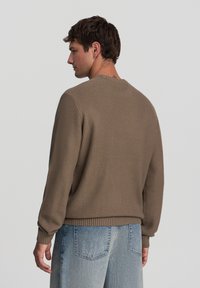 Maglione a maglia marrone con texture, con polsini e orlo a coste, caratterizzato da una vestibilità rilassata e scollo rotondo, abbinato a jeans di denim azzurri chiari.